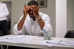 Photograph by Yan Krukau of a Black man sitting at a desk with a pencil in his hand and his hands on his head. He wears business casual attire, sits in an office setting, and appears to be very stressed.