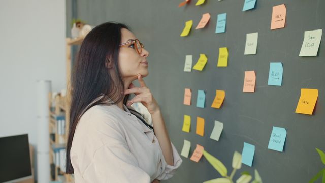 Thoughtful woman planning ideas on a chalkboard wall with colorful sticky notes in an office setting.